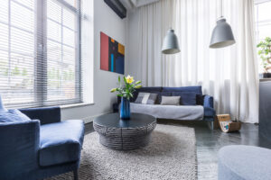 Living room with blue upholstered furniture, white faux-wood window blinds. gray industrial-chic hanging lamps and and white net curtain